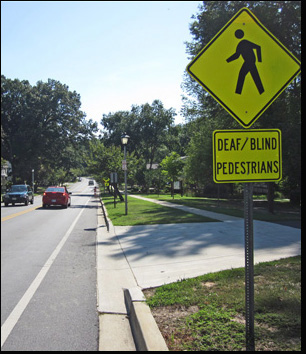 Both photos show the same crosswalk at a narrow two-lane residential street.  About 150 feet before the crosswalk we see a post with two signs on it - the top sign shows a drawing of a person walking, below that is a sign saying deaf/blind 'pedestrians.' Standing on the right side of the street is a woman wearing white shorts and a red-dand-white-checkered blouse, standing with one foot in front of the other and holding a white cane with the tip on the ground.  A red car has stopped just before reaching the sidewalk.